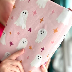 May include: A light pink fabric featuring a repeating pattern of white ghosts with black eyes and mouths, interspersed with small four-pointed stars in shades of orange, magenta, and light pink, creating a whimsical and festive textile design.