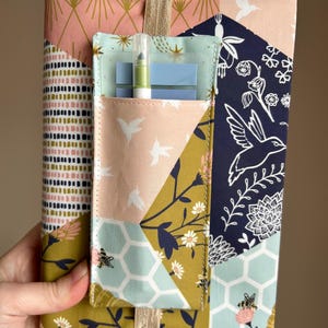 May include: A fabric journal cover with a patchwork design in pink, navy, olive green, and light blue. The cover features a pocket with a pen and sticky notes. The design includes hummingbirds, bees, and floral patterns. A gold ribbon is attached.