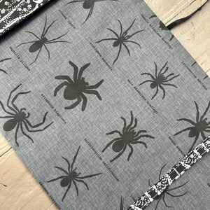 Puede incluir: Un rollo de tela gris con un patr&oacute;n repetitivo de ilustraciones de ara&ntilde;as negras, cada una etiquetada con su nombre cient&iacute;fico. El rollo est&aacute; asegurado con una correa blanca y negra decorada con un patr&oacute;n de calaveras y motivos florales.