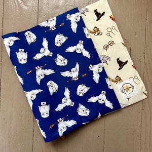 May include: A fabric book sleeve featuring a deep blue cover with a repeating pattern of white owls carrying letters and brooms. The inner layer is cream-colored with a print of wizarding hats, glasses, and wands, featuring a Mayberry Makers label.