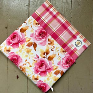 May include: A square fabric item featuring a white background with a pattern of pink roses and golden-brown leaves. One corner has a pink and white plaid section with a small white rectangular label that reads Mayberry Makes in black text.