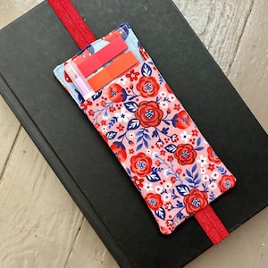 May include: A black book with a red elastic bookmark and a floral print pocket. The pocket is pink with red flowers, blue leaves, and white accents. Two sticky notes are visible inside the pocket.