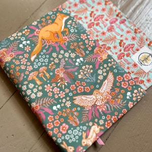 May include: A fabric item with a whimsical forest design. The pattern features an orange fox, owl, and rabbits amidst flowers and mushrooms in shades of green, pink, and orange. The item has a pink ribbon bookmark.