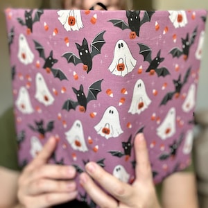 May include: A lavender fabric with a Halloween-themed pattern. The design features black bats, white ghosts holding jack-o'-lanterns, and candy corn scattered across the surface. The fabric is held by a person.