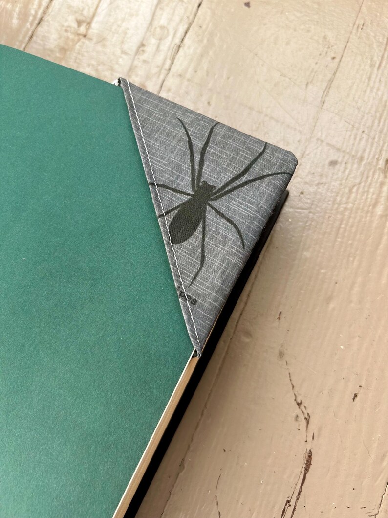 Puede incluir: Un marcap&aacute;ginas triangular de tela para la esquina de un libro con un patr&oacute;n gris texturizado y un gr&aacute;fico de ara&ntilde;a negra impreso en la superficie. El marcap&aacute;ginas est&aacute; sujeto a la esquina de una cubierta de libro verde liso con costuras blancas.