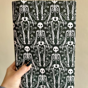 Puede incluir: Una pieza de tela rectangular con un patr&oacute;n repetitivo de esqueletos blancos sobre un fondo negro oscuro. Los esqueletos est&aacute;n intercalados con peque&ntilde;os motivos florales y de enredaderas blancas, creando un estampado gr&aacute;fico denso y continuo.