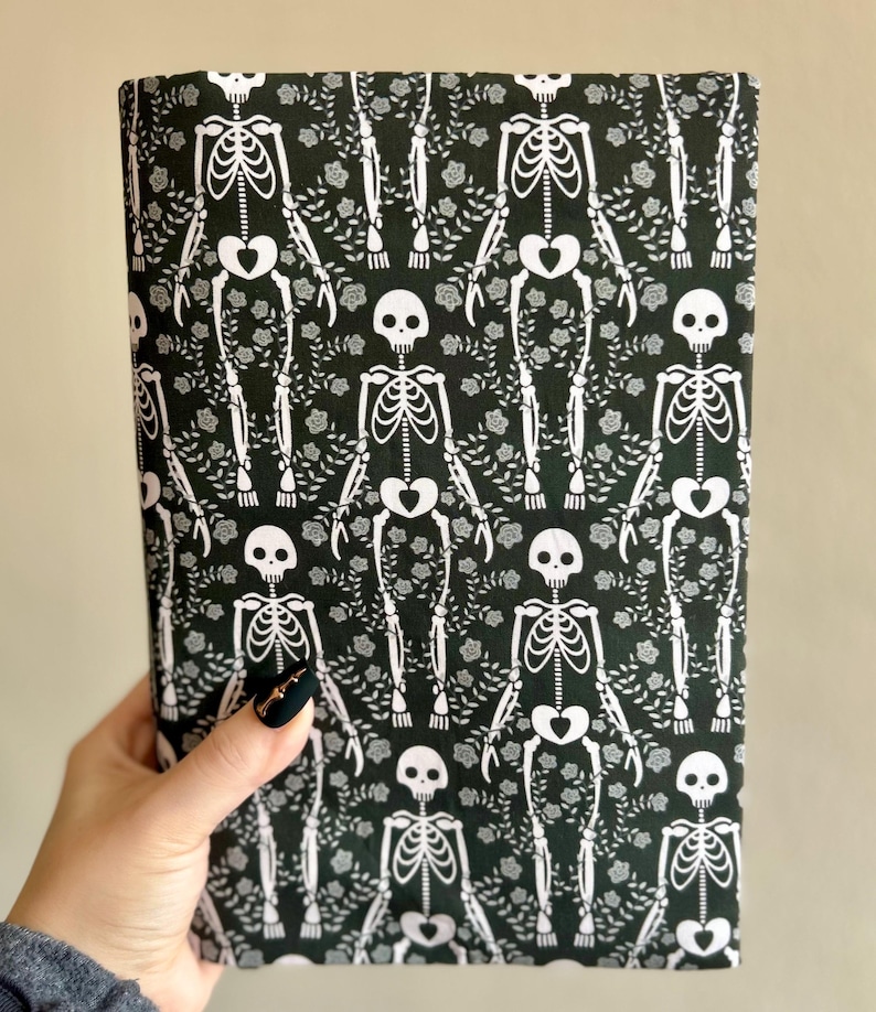 Puede incluir: Una pieza de tela rectangular con un patr&oacute;n repetitivo de esqueletos blancos sobre un fondo negro oscuro. Los esqueletos est&aacute;n intercalados con peque&ntilde;os motivos florales y de enredaderas blancas, creando un estampado gr&aacute;fico denso y continuo.