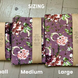 Puede incluir: Tres fundas de tela para libros en tama&ntilde;os peque&ntilde;o, mediano y grande, con fondo morado y un patr&oacute;n floral de rosas rosadas, hojas verdes y lunas crecientes y estrellas doradas. Cada funda tiene una banda de papel marr&oacute;n con texto.