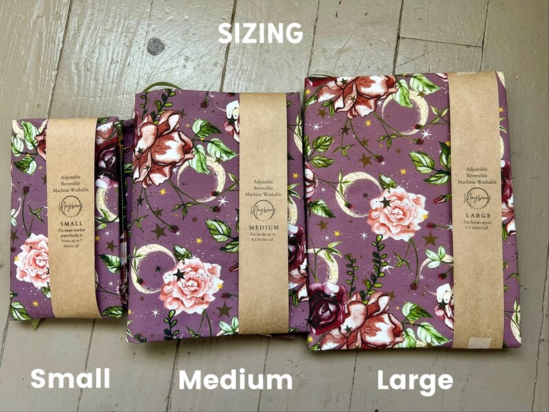 Puede incluir: Tres fundas de tela para libros en tama&ntilde;os peque&ntilde;o, mediano y grande, con fondo morado y un patr&oacute;n floral de rosas rosadas, hojas verdes y lunas crecientes y estrellas doradas. Cada funda tiene una banda de papel marr&oacute;n con texto.