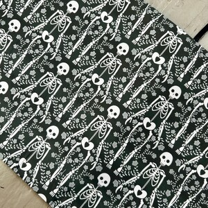 Puede incluir: Una tela negra con un patr&oacute;n blanco repetitivo de esqueletos con cajas tor&aacute;cicas en forma de coraz&oacute;n, rodeados de peque&ntilde;as enredaderas florales y hojas. El textil tiene un dise&ntilde;o estampado consistente en su superficie e incluye tiras negras.