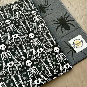 Puede incluir: Dos piezas de tela rectangulares. Una presenta un fondo negro con un patr&oacute;n de esqueletos blancos y flores. La otra es gris con ilustraciones de ara&ntilde;as negras, texto cient&iacute;fico y una etiqueta cuadrada blanca que dice Mayberry Makes.