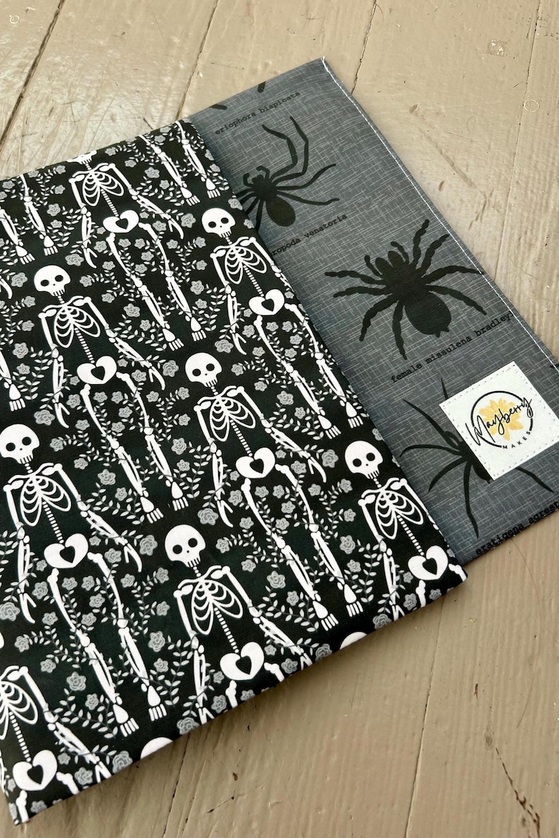 Puede incluir: Dos piezas de tela rectangulares. Una presenta un fondo negro con un patr&oacute;n de esqueletos blancos y flores. La otra es gris con ilustraciones de ara&ntilde;as negras, texto cient&iacute;fico y una etiqueta cuadrada blanca que dice Mayberry Makes.
