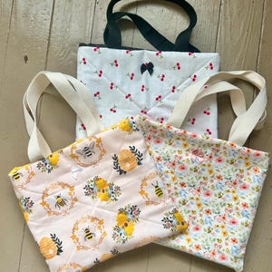 May include: Three quilted tote bags with different patterns. One bag has a white background with red cherries, another has a light pink background with bees and honeycombs, and the third has a floral pattern. All have fabric handles.