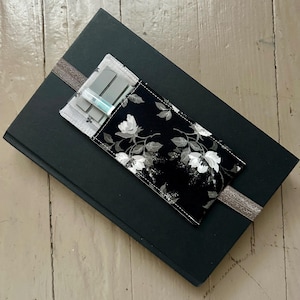May include: A black journal with a floral print pen holder. The pen holder is black with white floral designs. A silver ribbon is wrapped around the journal. The journal is on a light wood surface.