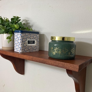 May include: A wooden wall shelf holding a potted green plant, a rectangular metal tin with a blue lid and blue and grey polka dots labeled PRAYER CARDS, and a dark green glass candle jar with a gold lid and gold leaf and acorn illustrations.