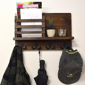May include: A dark brown wooden wall shelf with a built-in mail organizer and five hooks. The shelf has a small potted succulent plant and a glass jar on the top shelf. A plaid shirt, an umbrella, and a gray baseball cap are hanging on the hooks.