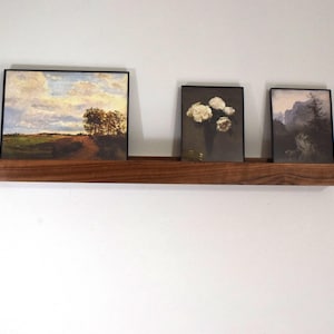 May include: A wooden shelf with three framed pictures. The pictures are of a landscape, a bouquet of white flowers, and a mountain scene.