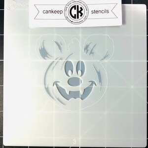 May include: A white plastic stencil of a Mickey Mouse pumpkin with a toothy grin. The stencil is on a gray grid background.