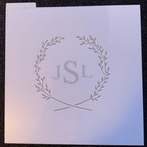 May include: A white stencil with a wreath design and the initials "JSL" in the center. The stencil is 4 inches in diameter.