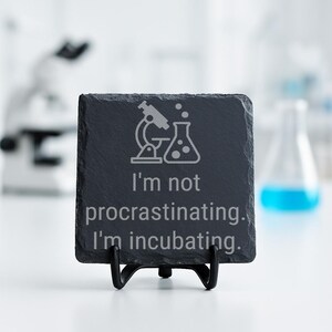 May include: A square, dark gray slate coaster with the text "I'm not procrastinating. I'm incubating." and a graphic of a microscope and beaker. The coaster is displayed on a black metal stand.