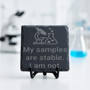 May include: A square, dark gray slate coaster with the text "My samples are stable. I am not." and a graphic of a microscope and a flask. The coaster is displayed on a black metal stand, with a blurred laboratory background.