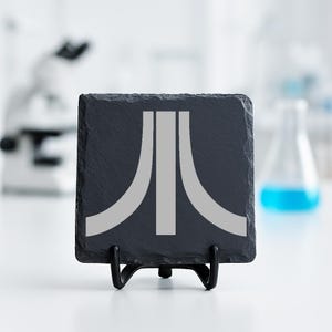 May include: A square, dark grey slate coaster featuring a light grey Atari logo. The coaster is presented on a black metal stand. The background is a blurred laboratory setting with scientific equipment.