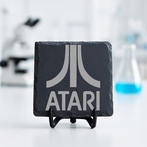 Op de afbeelding: Een vierkante, donkergrijze leistenen onderzetter met het Atari-logo in lichtgrijs. Het Atari-logo is een gestileerde bergketen met het woord "ATARI" eronder. De onderzetter wordt weergegeven op een zwarte metalen standaard.