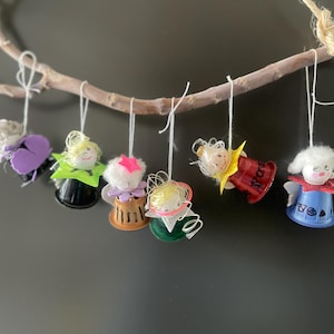 May include: A selection of handmade angel decorations suspended from a small branch. Each ornament has a unique design, with colourful details including purple, green, red, and blue. The angels have white hair and are hung by white string.