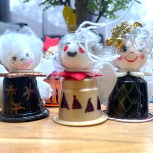 May include: Handmade angel ornaments. Each angel features a painted face, white hair, and fabric wings. The bodies are made from repurposed coffee pods, with decorative details. A festive, handcrafted item.