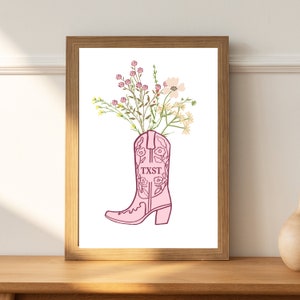 May include: A pink cowboy boot with floral embroidery and the word "TXST" on the side, filled with a bouquet of pink, white and yellow flowers.