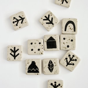 Op de afbeelding: Een set van tien witte keramische tegels met zwarte geometrische ontwerpen. De ontwerpen omvatten een sneeuwvlok, een boom, een huis, een blad, een tak, een berg, een boog en een reeks stippen.