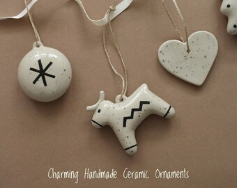 Puede incluir: Un conjunto de cinco adornos de cerámica hechos a mano con un esmalte blanco moteado y diseños pintados en negro. Los adornos incluyen una estrella, un corazón, un toro, un árbol y un círculo con una estrella en su interior. Los adornos están colgados de una cinta blanca y un cordel.
