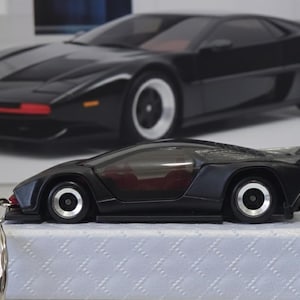 Op de afbeelding: Zwarte speelgoedauto met een strak design, zilveren velgen en een rode accent op de voorkant. Een grotere afbeelding van dezelfde auto staat op de achtergrond. De speelgoedauto staat op een wit oppervlak.