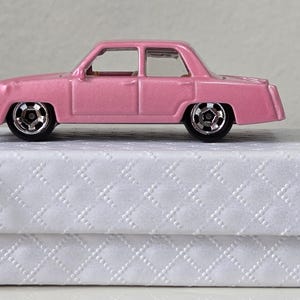 May include: A pink toy car with black wheels sits atop a white box with a quilted pattern. The car is a classic sedan style. A silver keychain is attached to the box. The overall theme is a miniature vehicle.