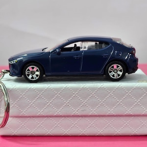 Puede incluir: Un llavero de coche de juguete azul oscuro en una caja blanca con un patrón de diamante. El coche tiene ruedas plateadas y un parabrisas transparente.