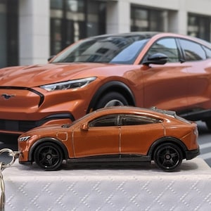Może przedstawiać: Pomarańczowy samochodzik Ford Mustang Mach-E z czarnymi kołami i brelokiem, przed pełnowymiarowym pomarańczowym Fordem Mustangiem Mach-E. Zabawkowy samochód stoi na białej powierzchni z rozmytym tłem miejskim.