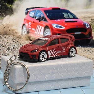Peut inclure: Un porte-clés de voiture jouet rouge avec des rayures blanches et un porte-clés argenté. Une plus grande voiture de rallye rouge est en arrière-plan, soulevant de la poussière. La voiture jouet est une réplique miniature d'une Ford Focus.