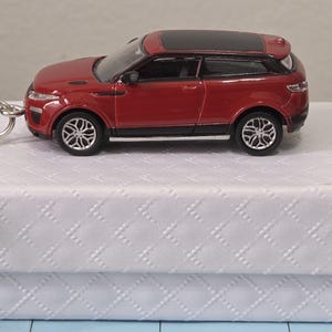 Könnte beinhalten: Ein roter Modellauto-Schlüsselanhänger mit schwarzem Dach und silbernen Rädern. Das Miniaturauto liegt auf einer weißen, strukturierten Box. Der Schlüsselanhänger hat einen silbernen Metallring.