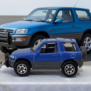 Könnte beinhalten: Ein blaues Spielzeugauto mit einem passenden SUV in Originalgröße im Hintergrund. Das Spielzeugauto ist an einem Schlüsselanhänger befestigt. Der SUV hat eine hellblaue Farbe. Das Spielzeugauto hat schwarze Reifen und einen grauen Boden.