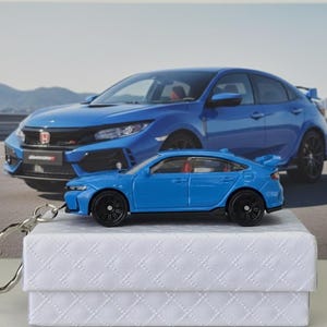 May include: A light blue die-cast model car with black wheels and a keychain. The model is on a white box, with a larger blue car image behind it. The car has a spoiler and red interior.