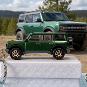 May include: A green die-cast model of a Ford Bronco SUV with gold-colored wheels and a keychain attachment. A larger green Bronco SUV is in the background. The keychain is attached to a white box with a diamond pattern.