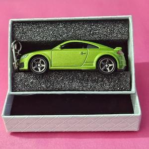 Puede incluir: Un llavero de coche de juguete verde en una caja blanca con interior negro. El coche es una réplica de un Audi TT.
