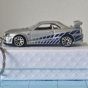 May include: Silver die-cast model car with blue and white graphics, a rear spoiler, and a keychain attachment. The car is displayed on a white, textured box. The car's wheels are black and silver.