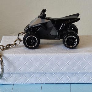Può includere: Un portachiavi ATV nero con una catena e un portachiavi argentati. Il veicolo fuoristrada in miniatura ha quattro ruote ed è appoggiato su una scatola bianca con un motivo a rombi.