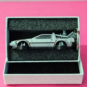 Könnte beinhalten: Ein silberner DeLorean Zeitmaschinen-Schlüsselanhänger in einer weißen Box mit schwarzem Schaumstoff-Innenraum.
