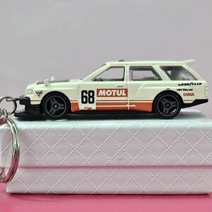 May include: White diecast model car with red and black accents. The car has the words "MOTUL" and "GOOD YEAR" on the side. The car is on a white box with a patterned design. The car has a key chain attached to it.