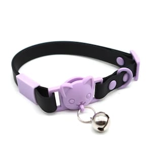 May include: A black and lavender cat collar with a lavender cat-shaped buckle and a silver bell. The collar is made of a black strap with a lavender buckle and a silver bell attached to the front.