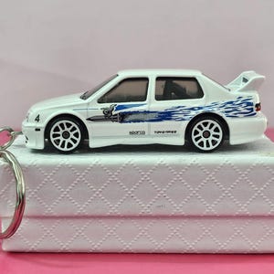 以下が含まれることがあります： 青い炎のデカールとリアスポイラーが付いた白いおもちゃの車。車は黒いホイールで、側面に4の数字があります。キーホルダーは白い箱に取り付けられています。
