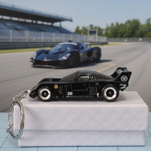 May include: A black toy race car with silver rims and a keychain attached. The car is on a white box, with a blurred race track and another black car in the background. The car has a spoiler and various decals.