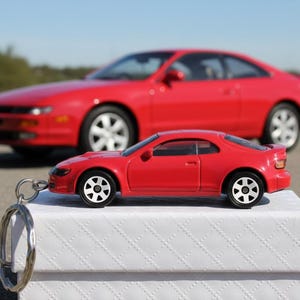 May include: A red toy car keychain with a matching full-size red car in the background. The keychain car is on a white box. The cars are on a paved surface with a blue sky in the background.